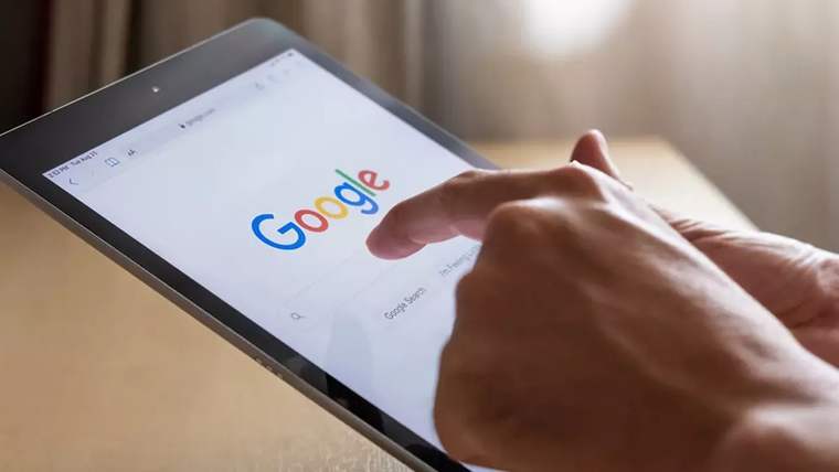 Masa üzerinde Google logosu ve arama sayfası açık bir tablete parmağıyla dokunan kişi