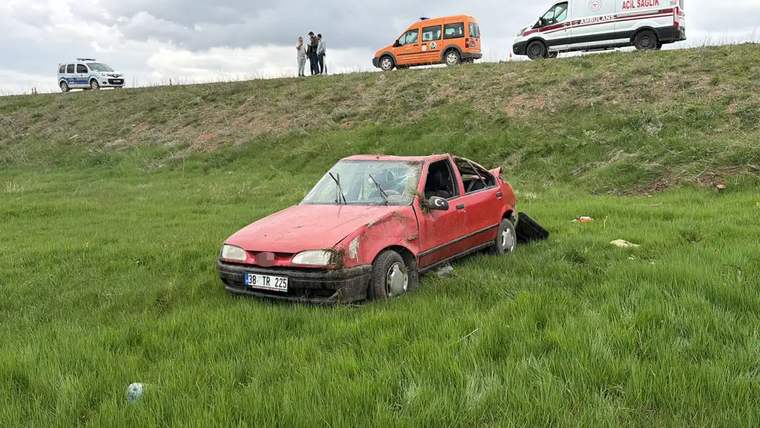 Kırmızı bir otomobil, şarampolde yan yatmış halde, etrafında yeşil çimenler var.
