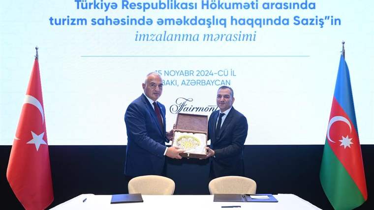 Türkiye ve Azerbaycan bayrakları önünde, iki kişi el sıkışarak anlaşma belgesi tutuyor.