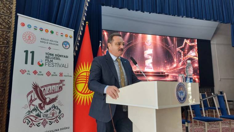 Kürsüde konuşan bir kişi, yanında Türk bayrağı ve Kırgızistan bayrağı, arkasında etkinlik afişi ve ekran bulunuyor.