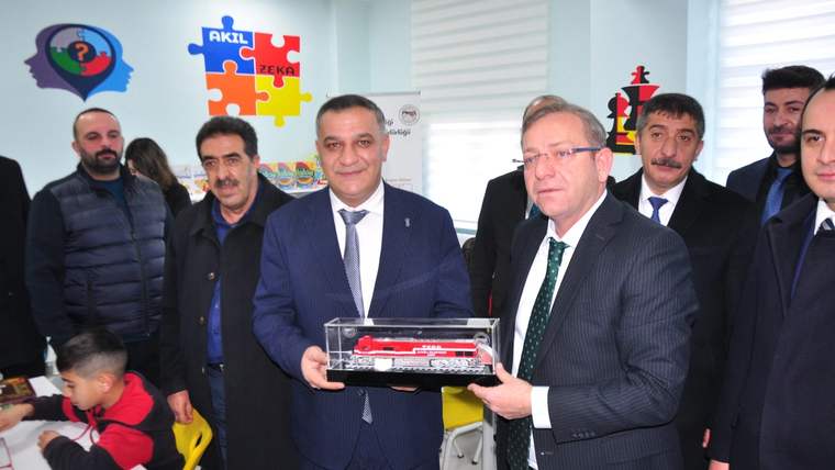 Bir grup erkek, yeni açılan sınıfta bir model tren tutarak poz veriyor. Arka planda akıl ve zeka oyunları temalı duvar süslemeleri var.