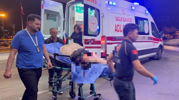 İki sağlık görevlisi, yüzü bulanıklaştırılmış bir kişiyi sedyeyle ambulansın önünde taşırken görülüyor.