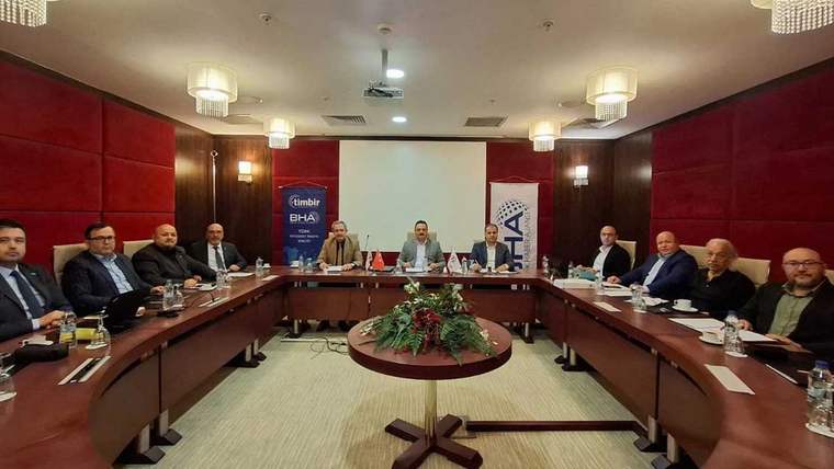 Bir toplantı odasında, uzun bir masa etrafında oturan dokuz kişi, belgeler ve içeceklerle birlikte görüntüleniyor.