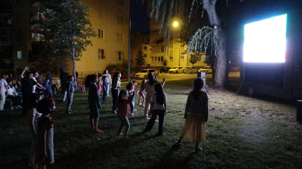 Gece, apartmanların önündeki çim alanda farklı yaşlardan insanlar aydınlatılmış ekranı izliyor.