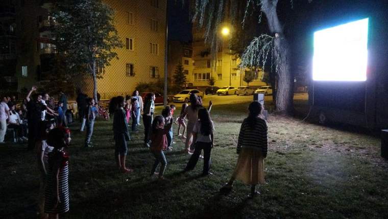 Gece, apartmanların önündeki çim alanda farklı yaşlardan insanlar aydınlatılmış ekranı izliyor.