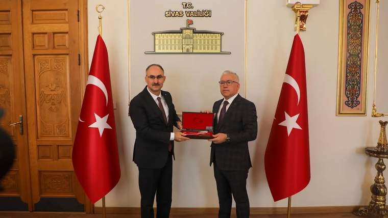İki takım elbiseli kişi, Türk bayrakları arasında kırmızı bir plaketi birlikte tutuyor.