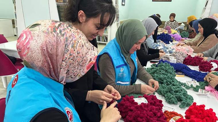 Kapalı bir salonda kadınlar, masadaki renkli yünleri elleriyle örerken görülüyor.