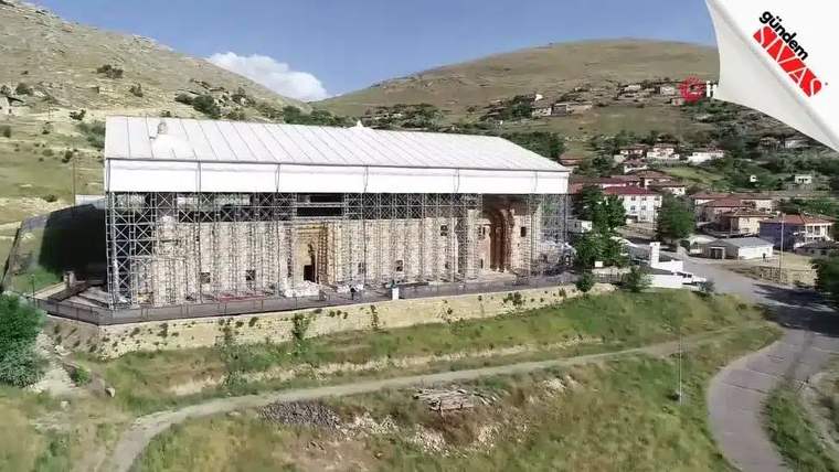 Dağ yamacındaki taş yapı, beyaz çatı ve metal iskelelerle çevrili; çevresinde evler ve kıvrımlı yol görülüyor.