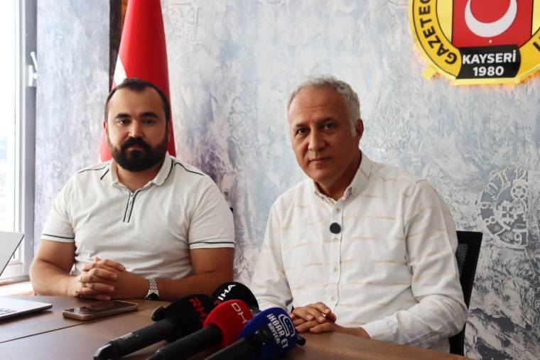 Masada yan yana oturan iki erkek, önlerindeki basın mikrofonları ve arkalarında Türk bayrağıyla görülüyor.