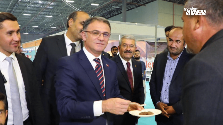 Fuar alanında takım elbiseli bir kişi, çevresindeki insanların önünde tabakta yiyecek taşırken görülüyor.