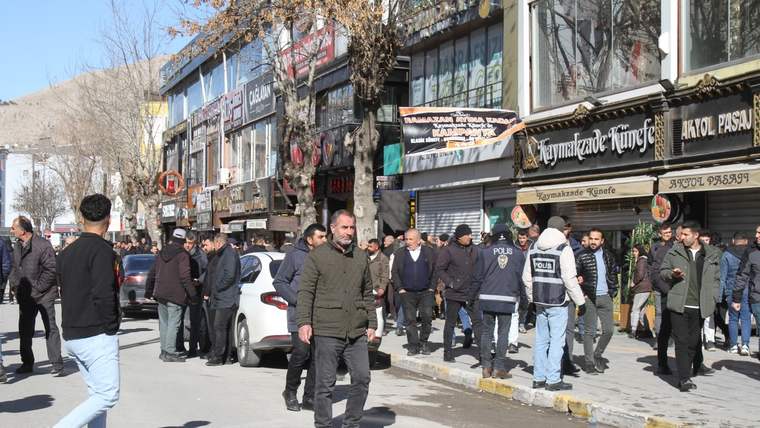 Mağazaların bulunduğu caddede polis yelekli görevliler ve kalabalık insanlar, park etmiş araçların yanında duruyor.