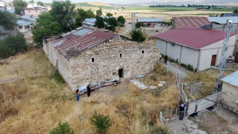 Taş duvarlı, çatısı kısmen çökmüş yapının çevresinde üç kişi ve kuru otlarla kaplı arazi görülüyor.