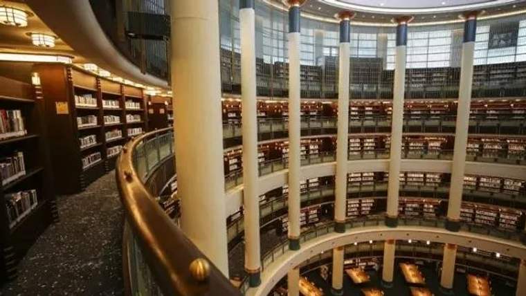 Çok katlı kütüphane salonunda kitap rafları, balkonlar ve yüksek pencereler görülüyor.