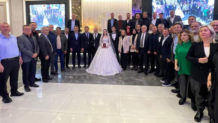 Gelin ve damat, salonda çevrelerindeki kalabalıkla birlikte kameraya karşı poz veriyor.