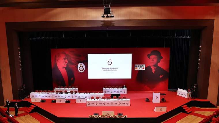 Kırmızı koltuklarla çevrili salonda, Galatasaray logolu ekran ve kürsülerin bulunduğu sahne görülüyor.