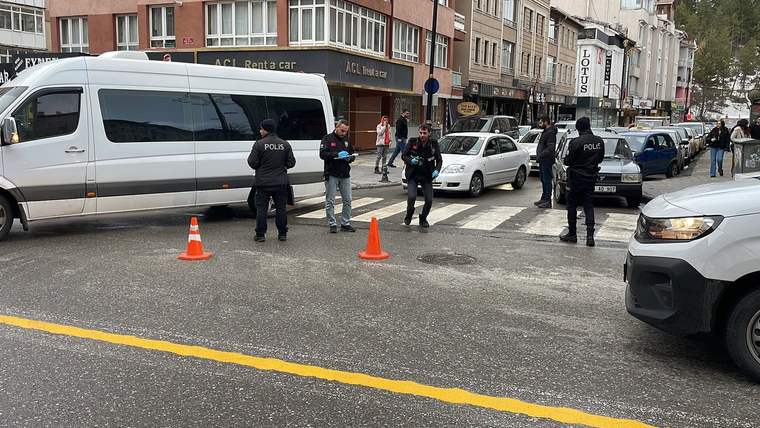 Üç polis memuru, turuncu trafik konileriyle çevrilen cadde üzerinde park etmiş araçların yanında duruyor.