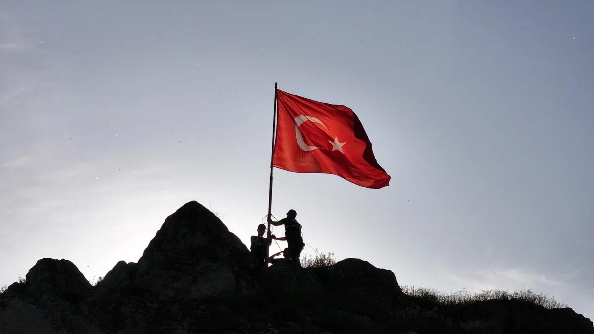 İki kişi, bayrağı bir tepe üzerinde dikmek için çalışıyor. Arkada Türk bayrağı dalgalanıyor.