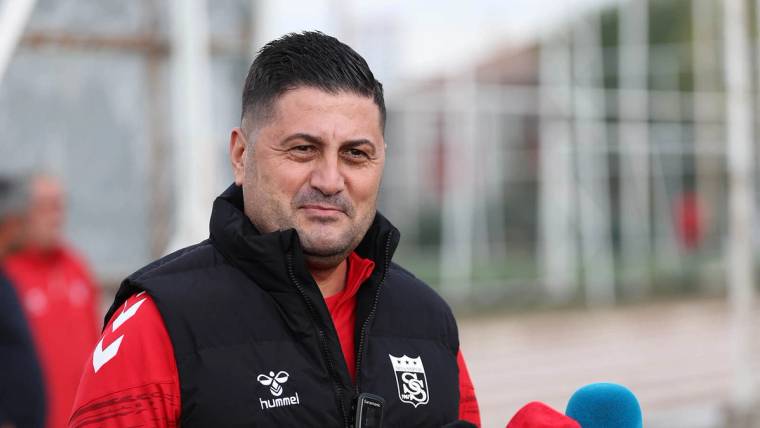 Spor kıyafeti ve yelek giyen bir kişi, önünde mavi mikrofonla dışarıda duruyor.
