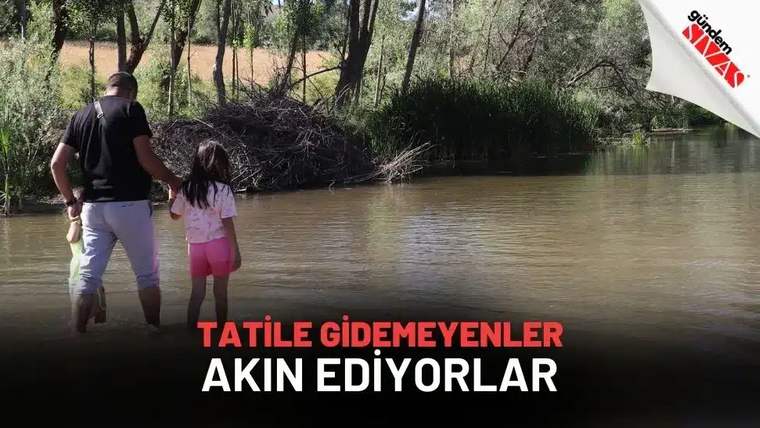 Bir adam ve küçük bir kız çocuğu, ağaçlık alandaki suyun kıyısında el ele yan yana duruyor.