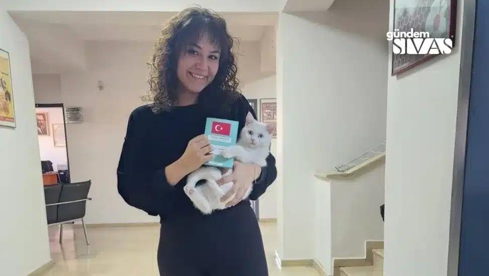 Bir kadın, kucağında beyaz bir kedi tutarak gülümsüyor ve elinde kimlik kartı tutuyor.