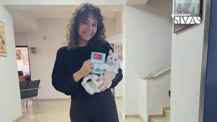 Bir kadın, kucağında beyaz bir kedi tutarak gülümsüyor ve elinde kimlik kartı tutuyor.
