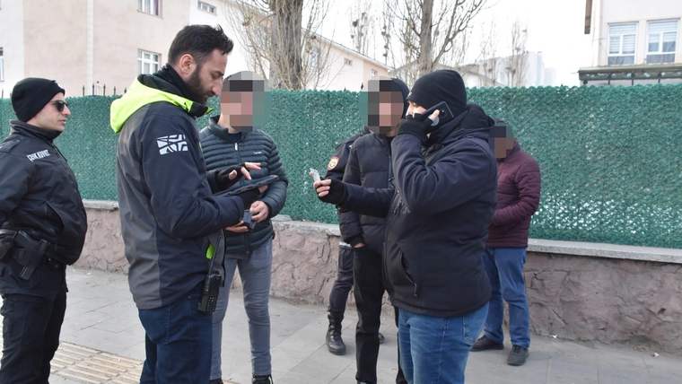 Bir okul binasının önündeki kaldırımda, montlu görevliler ve birkaç kişi birlikte duruyor.