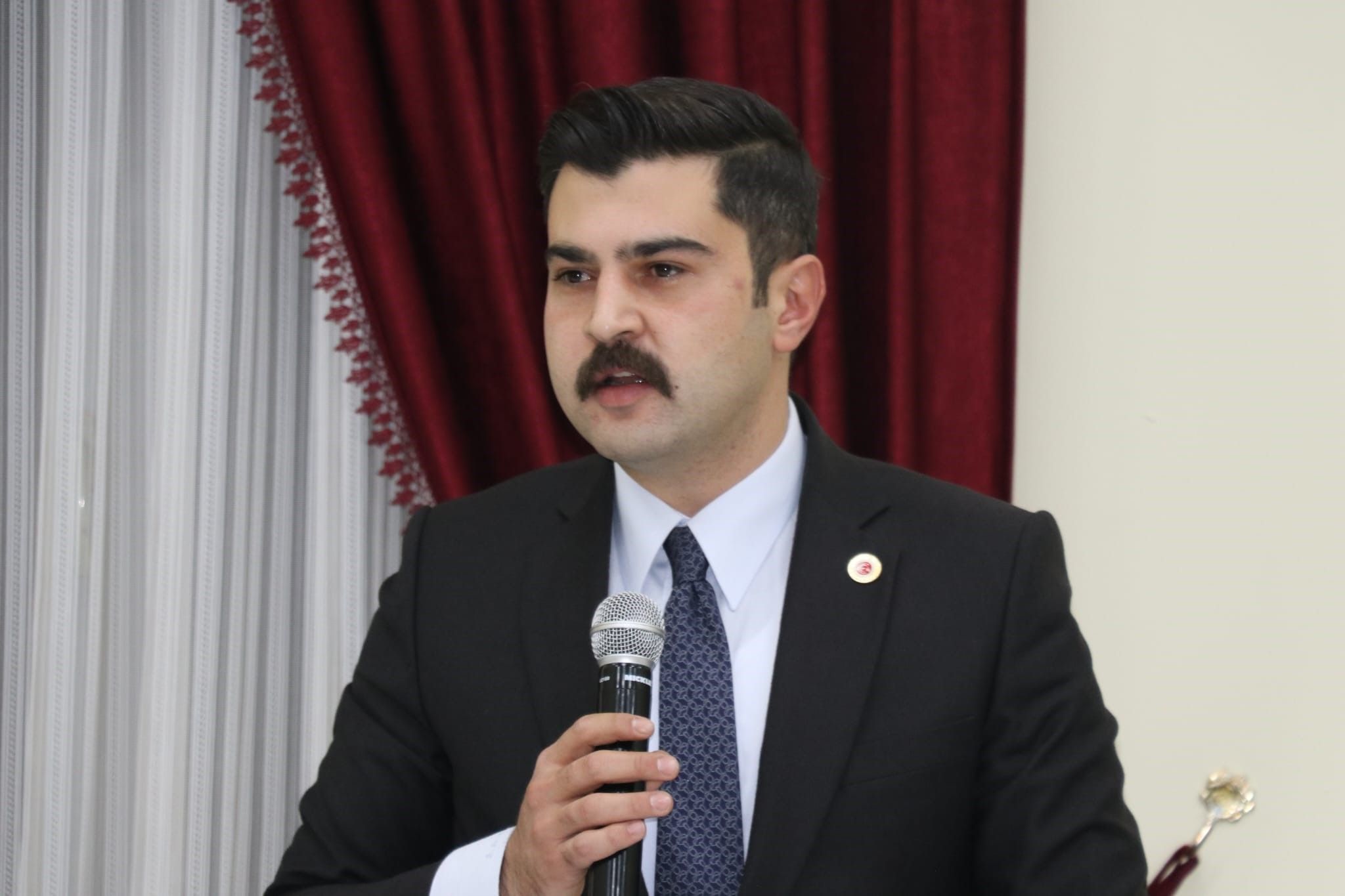 Kırmızı perdeli bir salonda mikrofonla konuşan takım elbiseli bir kişi.