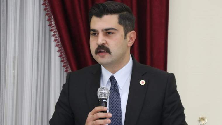 Kırmızı perdeli bir salonda mikrofonla konuşan takım elbiseli bir kişi.