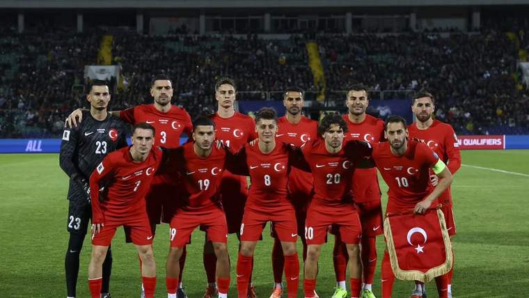 Yeşil sahada kırmızı formalar giyen 11 futbolcu yan yana poz verirken bir oyuncu Türk bayrağı tutuyor.