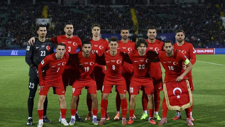 Yeşil sahada kırmızı formalar giyen 11 futbolcu yan yana poz verirken bir oyuncu Türk bayrağı tutuyor.