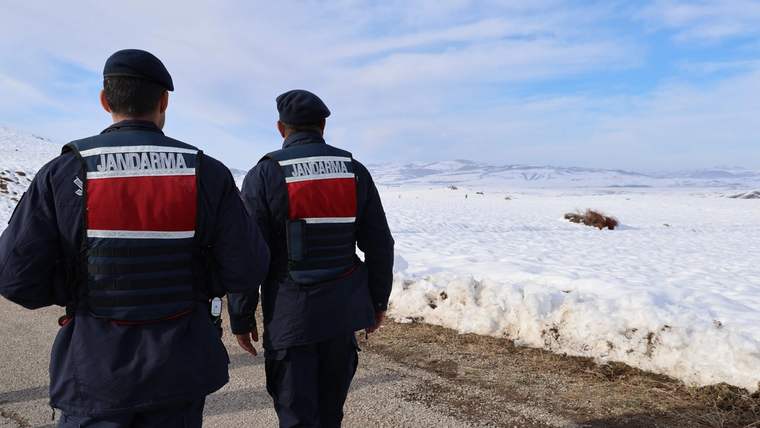 Karla kaplı arazide yürüyen iki jandarma, arka planda dağlar görünmekte.