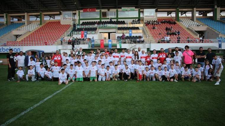 Stadyum sahasında çocuklar ve yetişkinlerden oluşan kalabalık bir grup, tribünlerin önünde toplu duruyor.
