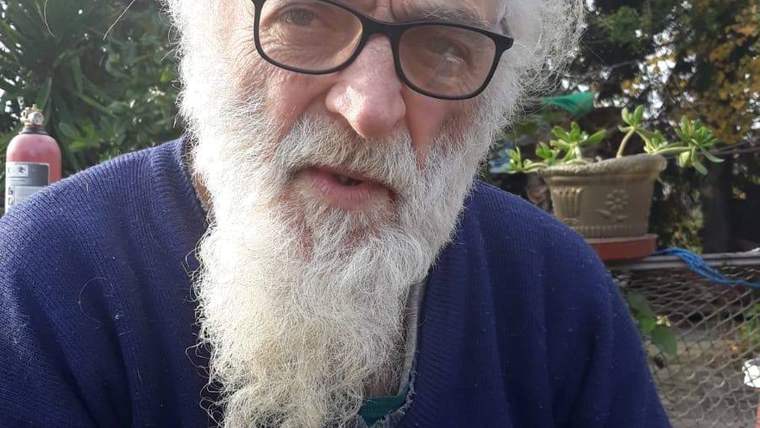 Beyaz sakallı bir adam, gözlük takarak kameraya bakıyor. Arka planda bitkiler ve bir yangın söndürücü var.