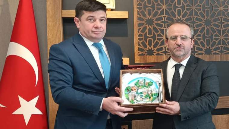 Azerbaycan bayrağı önünde duran iki erkek, üzerinde renkli çizim bulunan ahşap plaketi birlikte tutuyor.