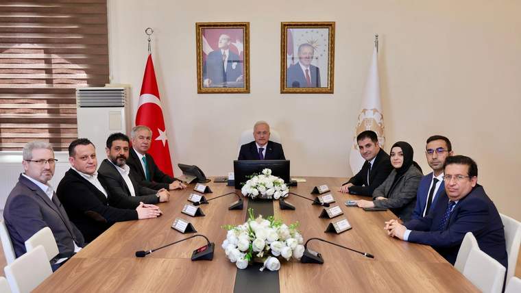 Toplantı masasında oturan dokuz kişi, Türk bayrağı ve başka bir bayrakla çevrili salonda görülüyor.