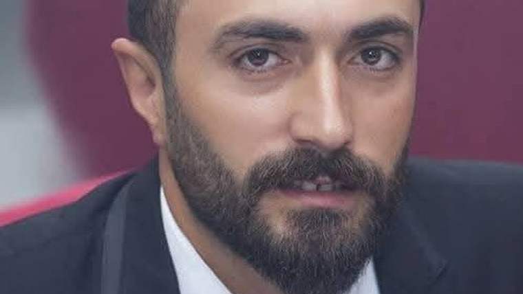Kareli gömlek giymiş, sakallı bir adamın yüzü yakın planda. Arka planda bulanık bir alan var.