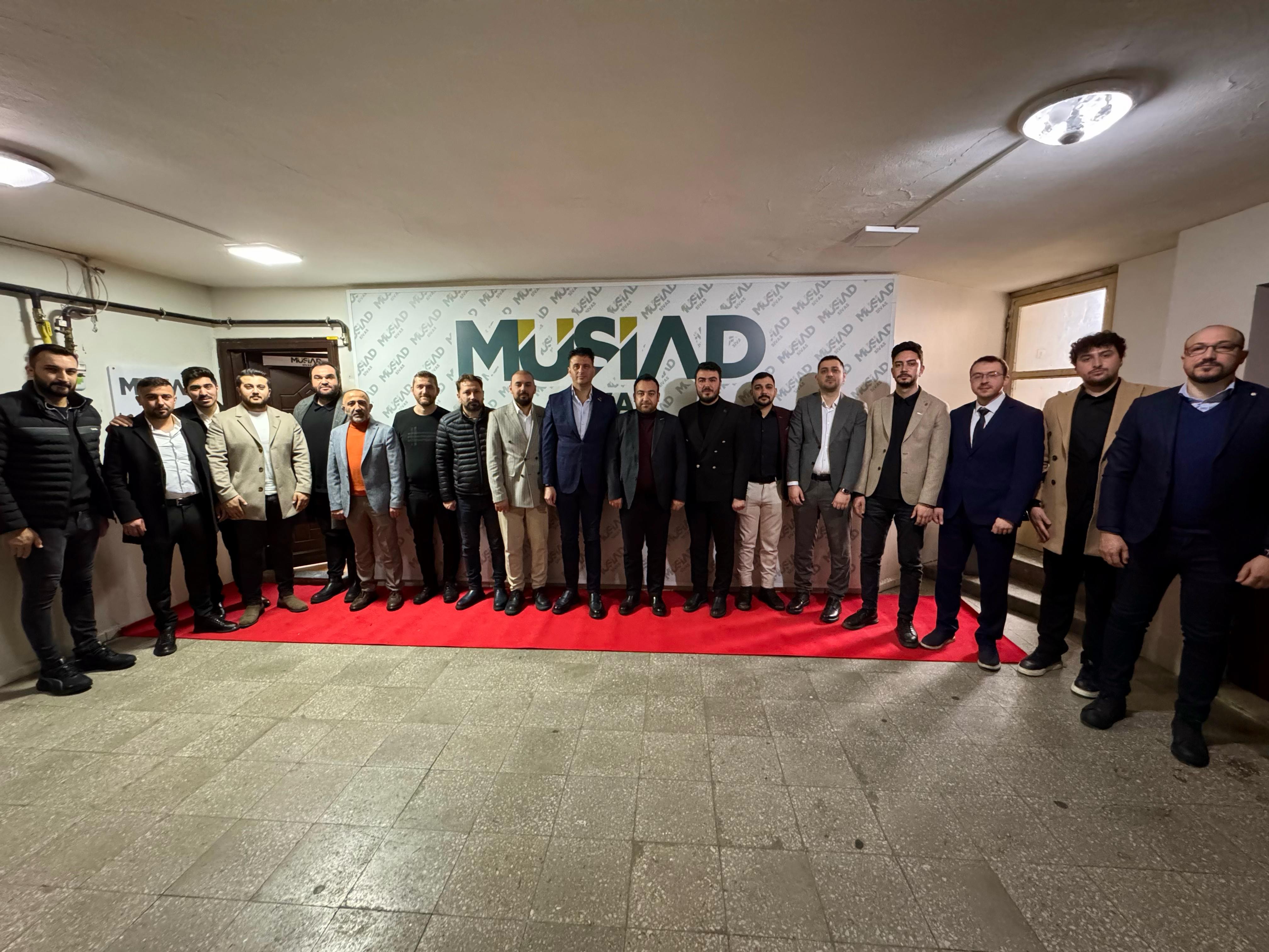 MÜSİAD yazılı pankartın önünde, kapalı salonda yan yana duran bir grup erkek