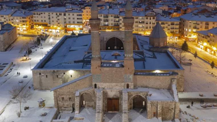 Karla kaplı bir alanda, iki minaresi olan tarihi bir cami ve çevresindeki binalar görünüyor.