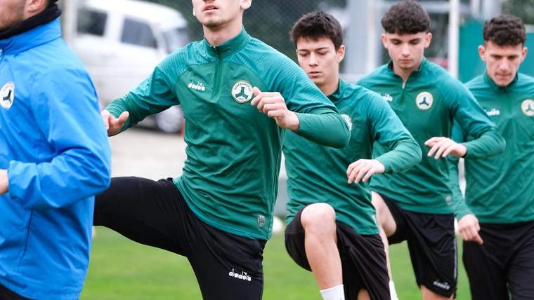 Yeşil eşofmanlı futbolcular, çim sahada antrenman yaparken bacaklarını kaldırıyor; önde mavi montlu biri bulunuyor.