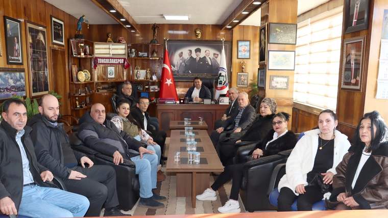 Ahşap mobilyalı bir odada, Türk bayrakları ve duvar süsleri önünde oturan bir grup kişi görülüyor.