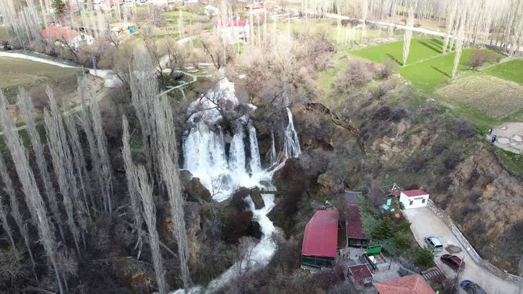 Sızır Şelalesi'nden akan su, çevresindeki ağaçlar ve yapılarla birlikte görünmekte.