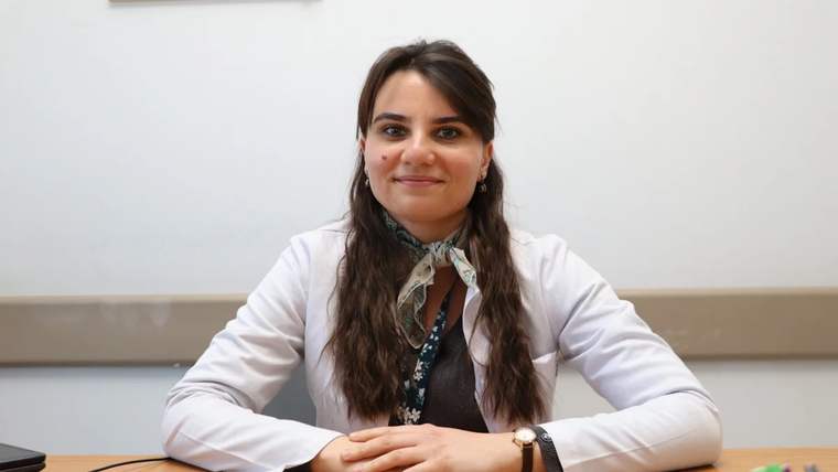 Beyaz önlüklü bir kadın, masa başında ellerini birleştirerek oturuyor; yanında kalemlik bulunuyor.
