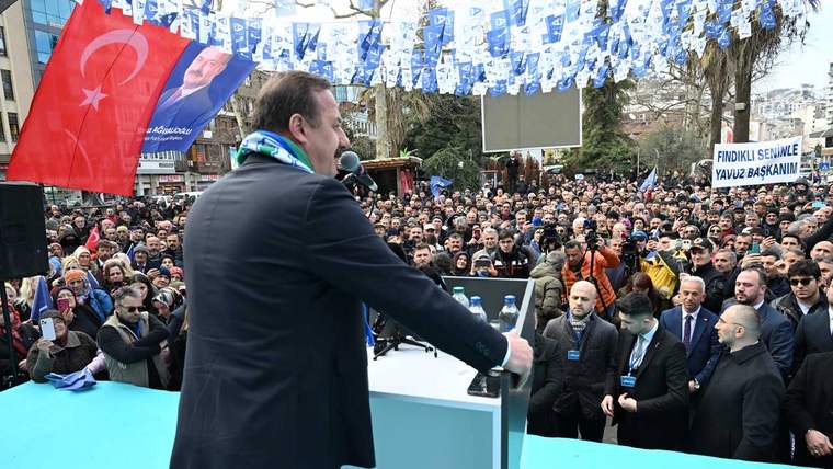 Kürsüde konuşan bir kişi, mavi-beyaz bayraklarla süslenmiş alandaki kalabalığa sesleniyor.