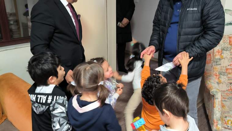 İç mekânda iki yetişkin erkeğin çevresinde toplanan çocuklardan biri, elindeki kâğıdı uzatıyor.