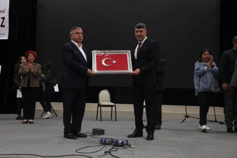 Sahnede iki erkek, çerçeveli Türk bayrağını birlikte tutarken arkada izleyiciler ve mikrofon görülüyor.