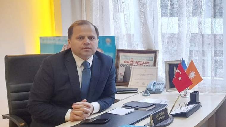 Takım elbiseli bir kişi, üzerinde iki bayrak ve telefon bulunan ofis masasının yanında oturuyor.