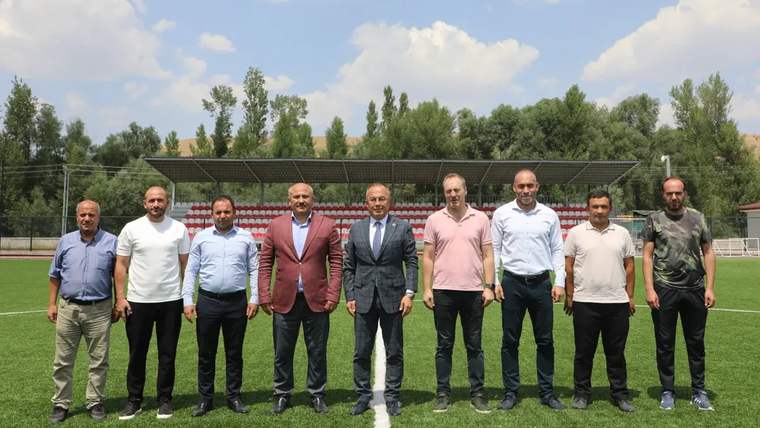 Saha denetimlerinde bulunan bir grup erkek, futbol sahasında yan yana duruyor.