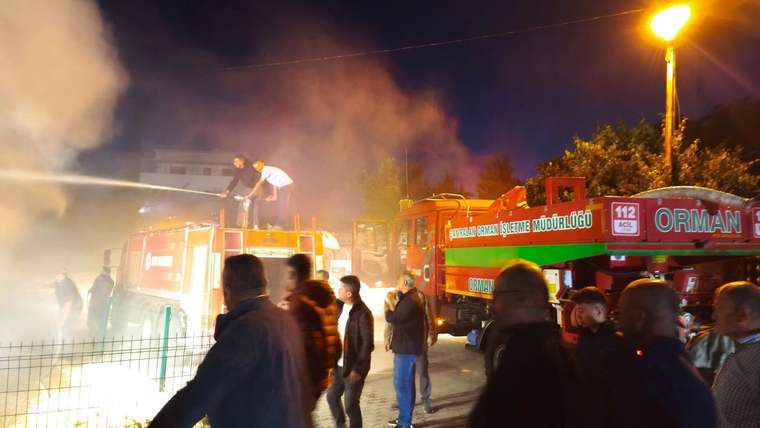 Gece saatlerinde alev ve dumanların yükseldiği alanda itfaiye araçları ve izleyen insanlar görülüyor.