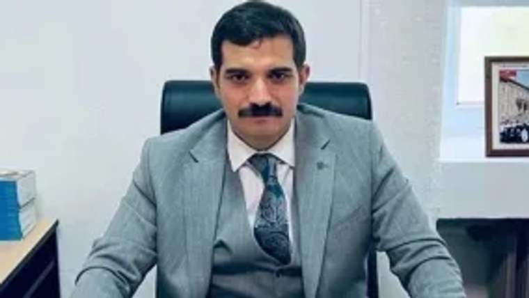Masada oturan bir kişi, gri takım elbise giymekte ve ciddi bir ifadeye sahip.