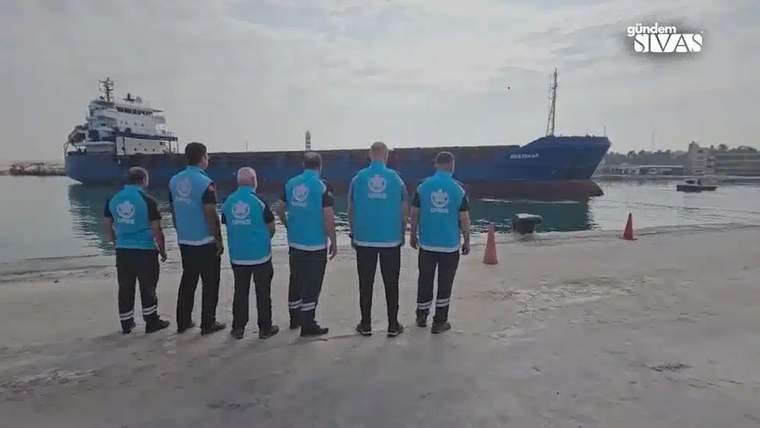 Mavi yelek giymiş altı kişi, bir geminin yanaştığı limanda duruyor.